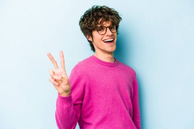 Mavi arka planda izole edilmiş genç beyaz adam zafer işareti gösteriyor ve gülümsüyor..