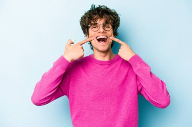 Mavi arka planda izole edilmiş genç beyaz adam gülümsüyor, parmaklarını ağıza doğrultuyor..