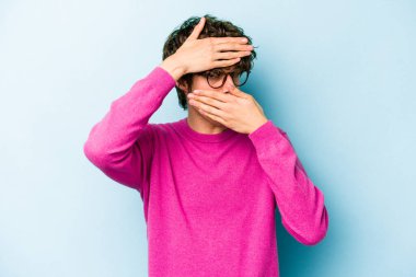 Mavi arka planda izole edilmiş genç beyaz adam kameraya parmaklarıyla göz kırpıyor, yüzünü kapatmaktan utanıyor..