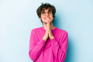 Mavi arka planda izole edilmiş genç beyaz bir adam el ele tutuşup, ağzının kenarında dua ediyor, kendinden emin..