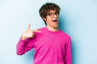 Mavi arka planda izole edilmiş genç beyaz adam bir gömlek fotokopi alanını işaret ediyor, gururlu ve kendinden emin.