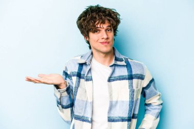 Kafkasyalı genç adam mavi arka planda izole edilmiş sorgulama jestinde şüphe ediyor ve omuz silkiyor..