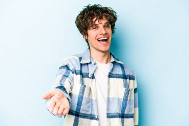 Mavi arka planda izole edilmiş genç beyaz adam selamlama jesti olarak kameraya elini uzatıyor..