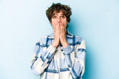 Mavi arka planda izole edilmiş genç beyaz adam elleriyle ağzını kapatırken şok oldu..