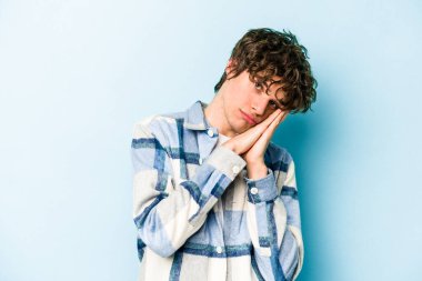 Mavi arka planda izole edilmiş genç beyaz adam esniyor. Ağzı eliyle kapatan yorgun bir hareket sergiliyor..