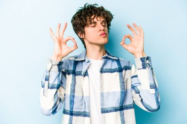 Mavi arka planda izole edilmiş genç beyaz adam yorucu bir günün ardından rahatlıyor. Yoga yapıyor..