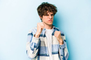 Mavi arka planda izole edilmiş genç beyaz adam yumruğunu kameraya gösteriyor, agresif yüz ifadesi var..