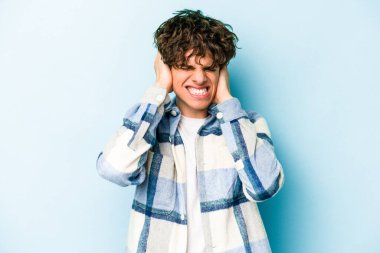 Mavi arka planda izole edilmiş genç beyaz adam kulaklarını elleriyle kapatıyor..