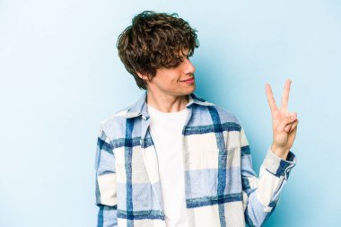 Kafkasyalı genç adam mavi arka planda izole edilmiş neşeli ve kaygısız parmaklarıyla barış sembolü gösteriyor..
