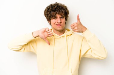 Beyaz arka planda izole edilmiş genç beyaz bir adam baş parmaklarını aşağı ve yukarı kaldırıyor, zor seçim konsepti