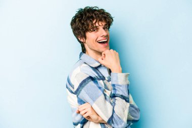 Mavi arka planda izole edilmiş genç beyaz adam gülümsüyor ve kendinden emin, elleriyle çenesine dokunuyor..