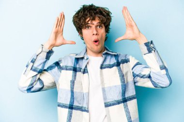 Mavi arka planda izole edilmiş genç beyaz bir adam zaferi ya da başarıyı kutluyor, şaşırıyor ve şok oluyor..