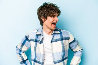 Mavi arka planda izole edilmiş genç beyaz adam mutlu bir şekilde gülüyor ve ellerini karnının üstünde tutarak eğleniyor..