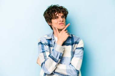 Mavi arka planda izole edilmiş genç beyaz adam alaycı bir ifadeyle kameraya bakarken mutsuz oluyor..