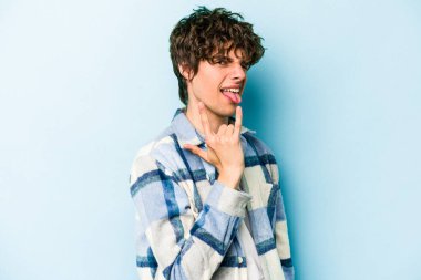 Mavi arka planda izole edilmiş genç beyaz adam parmaklarıyla kaya hareketi gösteriyor.