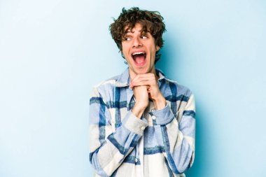 Mavi arka planda izole edilmiş genç beyaz adam ellerini çenesinin altında tutuyor, mutlu bir şekilde yana bakıyor..