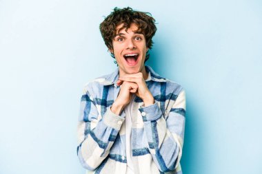 Kafkasyalı genç adam mavi arka planda izole edilmiş şans için dua ediyor, şaşırmış ve ağzını açıp önüne bakıyor..