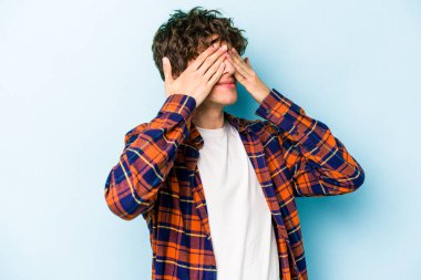 Mavi arka planda izole edilmiş genç beyaz adam elleriyle gözlerini kapatmaktan korkuyor..