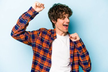 Mavi arka planda izole genç beyaz adam özel bir günü kutluyor, atlıyor ve enerjiyle kollarını kaldırıyor..