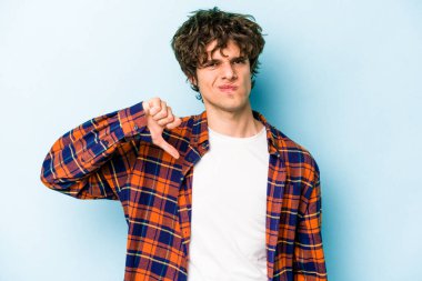 Mavi arka planda izole genç beyaz adam baş parmağını aşağı gösteriyor, hayal kırıklığı konsepti.