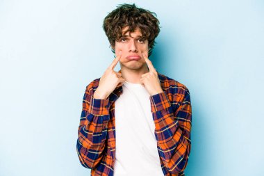 Kafkasyalı genç adam mavi arka planda izole edilmiş, ağlıyor, bir şeyden memnun değil, acı ve kafa karışıklığı konsepti..