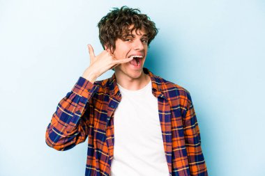 Mavi arka planda izole edilmiş genç beyaz adam parmaklarıyla bir cep telefonu hareketi gösteriyor..