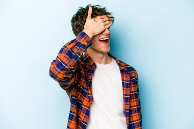 Mavi arka planda soyutlanmış genç beyaz bir adam elleriyle gözlerini kapatıyor, geniş bir gülümsemeyle bir sürpriz bekliyor..