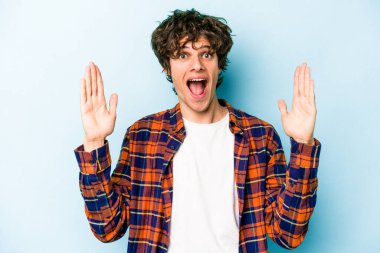 Mavi arka planda izole edilmiş genç beyaz adam hoş bir sürpriz alıyor, heyecanlı ve el kaldırıyor..