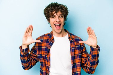 Kafkasyalı genç adam mavi arka planda izole edilmiş. Elinde avuç içi olan bir şey var ve kameraya sunuyor..