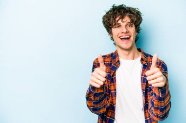Mavi arka planda izole edilmiş genç beyaz adam gülümsüyor ve başparmağını kaldırıyor.