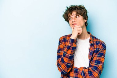 Mavi arka planda izole edilmiş genç beyaz adam şüpheli ve şüpheci bir ifadeyle yana bakıyor..