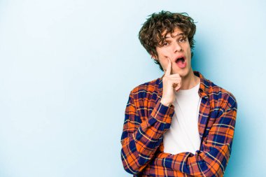 Mavi arka planda izole edilmiş genç beyaz adam şüpheli ve şüpheci bir ifadeyle yana bakıyor..