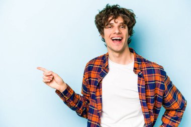 Mavi arka planda izole edilmiş genç beyaz adam işaret parmağıyla işaret ederek gülümsüyor..