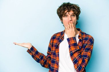 Mavi arka planda izole edilmiş genç beyaz adam, avuç içi fotokopi alanını elinde tutmaktan etkilenmiş..