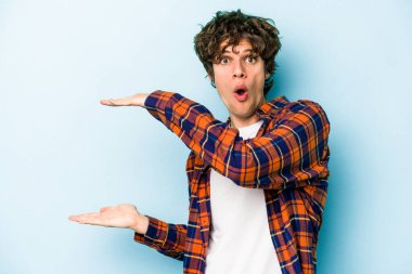 Mavi arka planda izole genç beyaz adam şok ve hayret içinde elleri arasında bir kopya boşluk tutuyor.