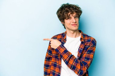 Mavi arka planda izole edilmiş genç beyaz adam gülümsüyor ve boş alanda bir şeyler gösteriyor..