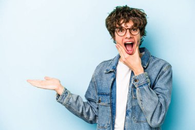 Mavi arka planda izole edilmiş genç beyaz bir adam avucunun içinde fotokopi alanı tutuyor, el üstünde tutuyor. Şaşırmış ve mutlu..