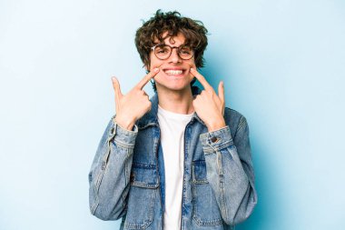 Mavi arka planda izole edilmiş genç beyaz adam gülümsüyor, parmaklarını ağıza doğrultuyor..