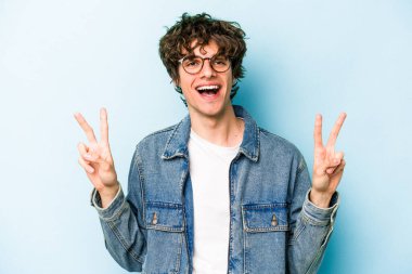 Mavi arka planda izole edilmiş genç beyaz adam zafer işareti gösteriyor ve gülümsüyor..