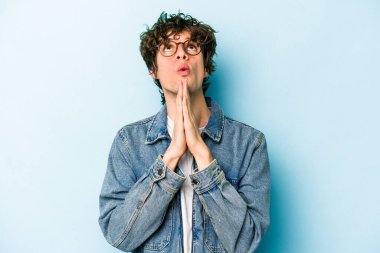Mavi arka planda izole edilmiş genç beyaz bir adam el ele tutuşup, ağzının kenarında dua ediyor, kendinden emin..