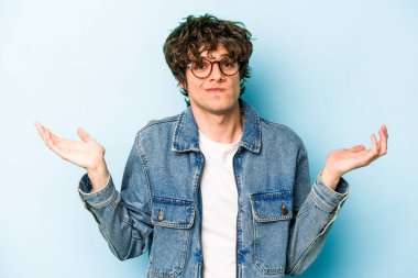 Kafkasyalı genç adam mavi arka planda izole edilmiş sorgulama jestinde şüphe ediyor ve omuz silkiyor..