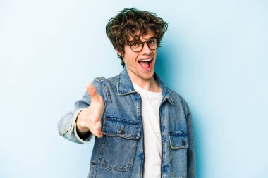 Mavi arka planda izole edilmiş genç beyaz adam selamlama jesti olarak kameraya elini uzatıyor..