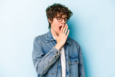 Mavi arka planda izole edilmiş genç beyaz adam esniyor. Ağzı eliyle kapatan yorgun bir hareket sergiliyor..