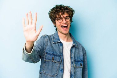 Mavi arka planda izole edilmiş genç beyaz adam gülümseyerek beş numarayı parmaklarıyla gösteriyor..