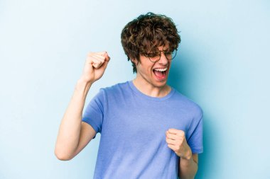 Kafkasyalı genç adam mavi arka planda izole edilmiş kaygısız ve heyecanlı tezahüratlar yapıyor. Zafer konsepti.