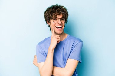 Mavi arka planda izole edilmiş genç beyaz adam gülümsüyor ve kendinden emin, elleriyle çenesine dokunuyor..