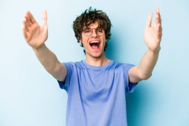 Mavi arka planda izole edilmiş genç beyaz bir adam zaferi ya da başarıyı kutluyor, şaşırıyor ve şok oluyor..