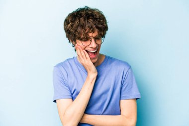 Mavi arka planda izole edilmiş genç beyaz adam mutlu bir şekilde gülüyor ve ellerini karnının üstünde tutarak eğleniyor..