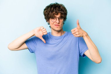 Kafkasyalı genç adam mavi arka planda izole edilmiş, baş parmağını kaldırıyor, zor seçim konsepti.