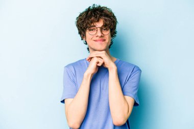 Mavi arka planda izole edilmiş genç beyaz adam ellerini çenesinin altında tutuyor, mutlu bir şekilde yana bakıyor..